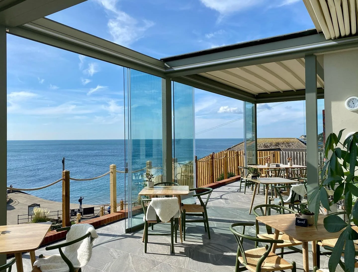Indoor view of a seaside cafe with glass walls, wooden tables, chairs, and potted plants, overlooking the ocean under a blue sky with clouds.