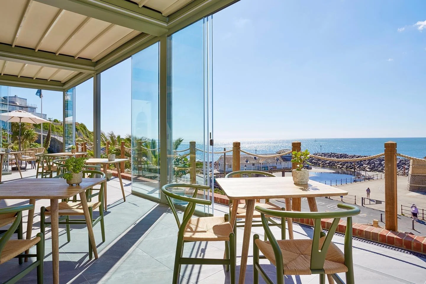 Sunlit indoor seating with wooden tables and green chairs overlooking a beach and ocean through large glass windows.