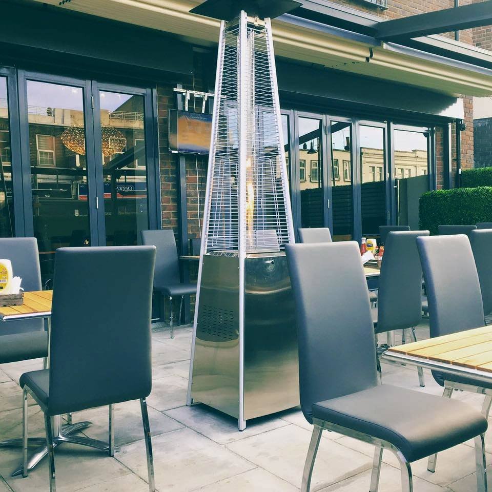 Outdoor patio with gray chairs and tables, and a tall stainless steel patio heater, in front of large glass doors and windows.