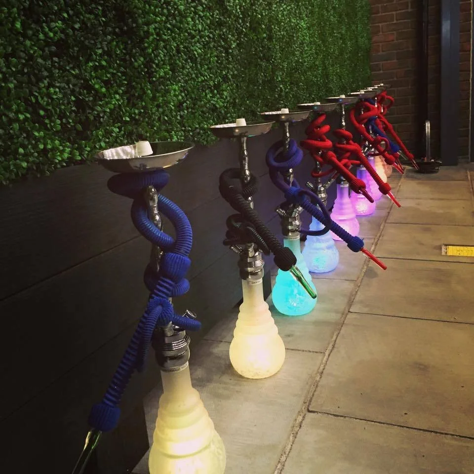 Six illuminated hookah pipes with glowing bases and colorful hoses, lined up on a sidewalk against a brick wall and green shrubbery.