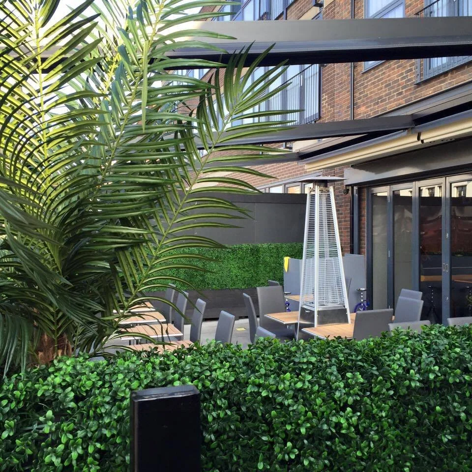 Outdoor patio area with a tall patio heater, several tables and chairs, surrounded by green bushes and a tall palm plant, with a building in the background.