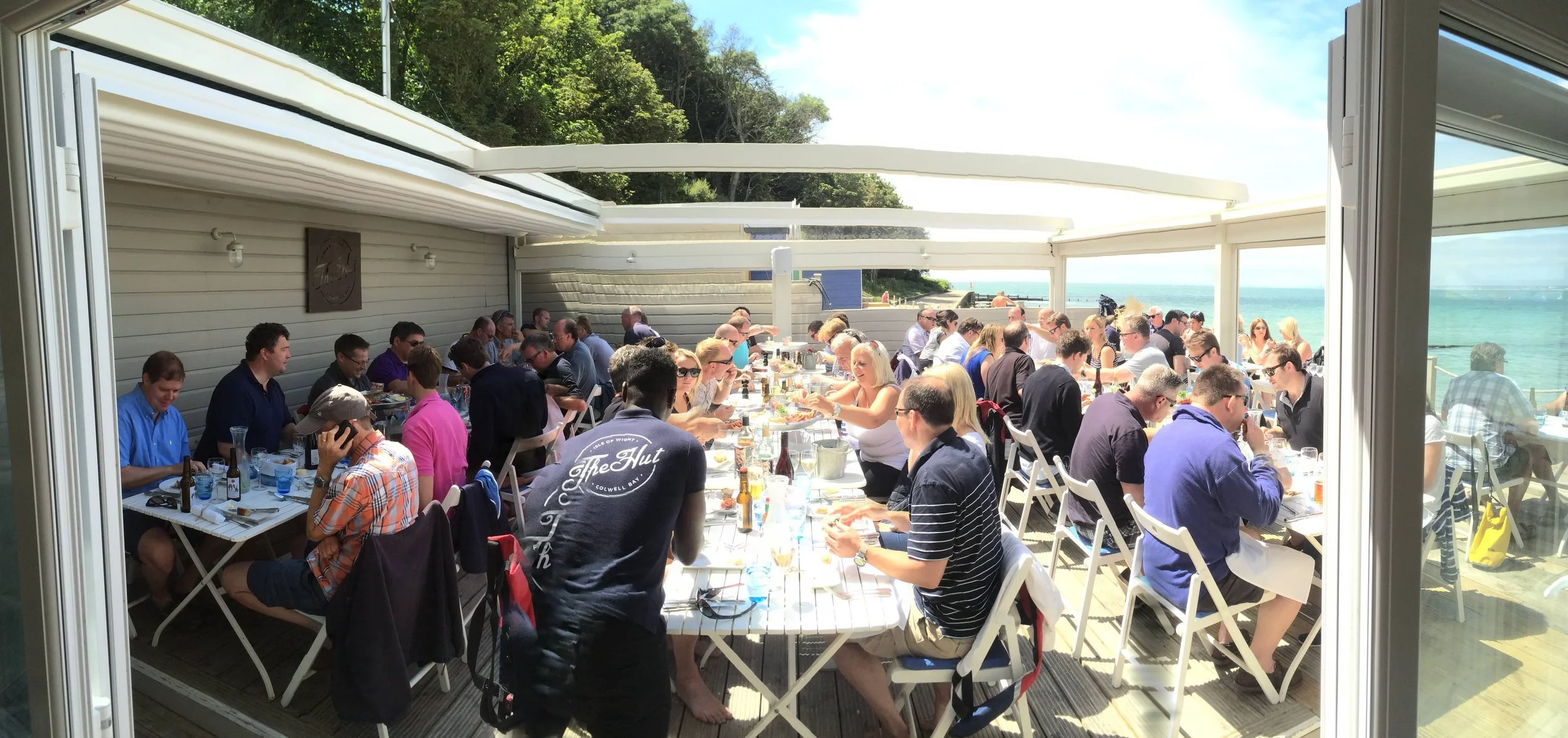 A large outdoor gathering of people dining at a restaurant terrace overlooking the beach, with tables filled with food and drinks and the ocean in the background.
