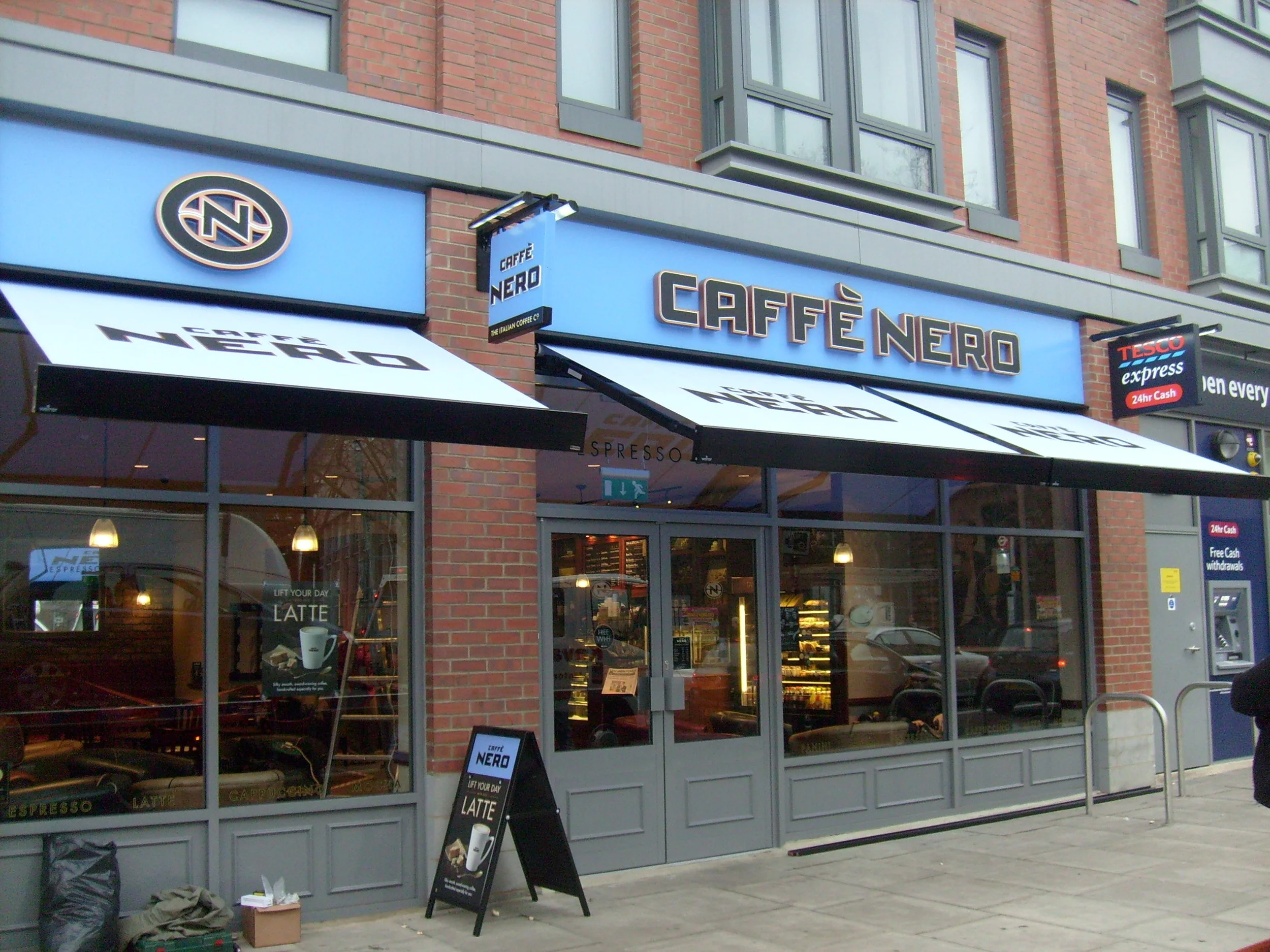 Exterior of Caffè Nero coffee shop with blue signage and black and white awnings, large glass windows, and a sidewalk display sign outside.