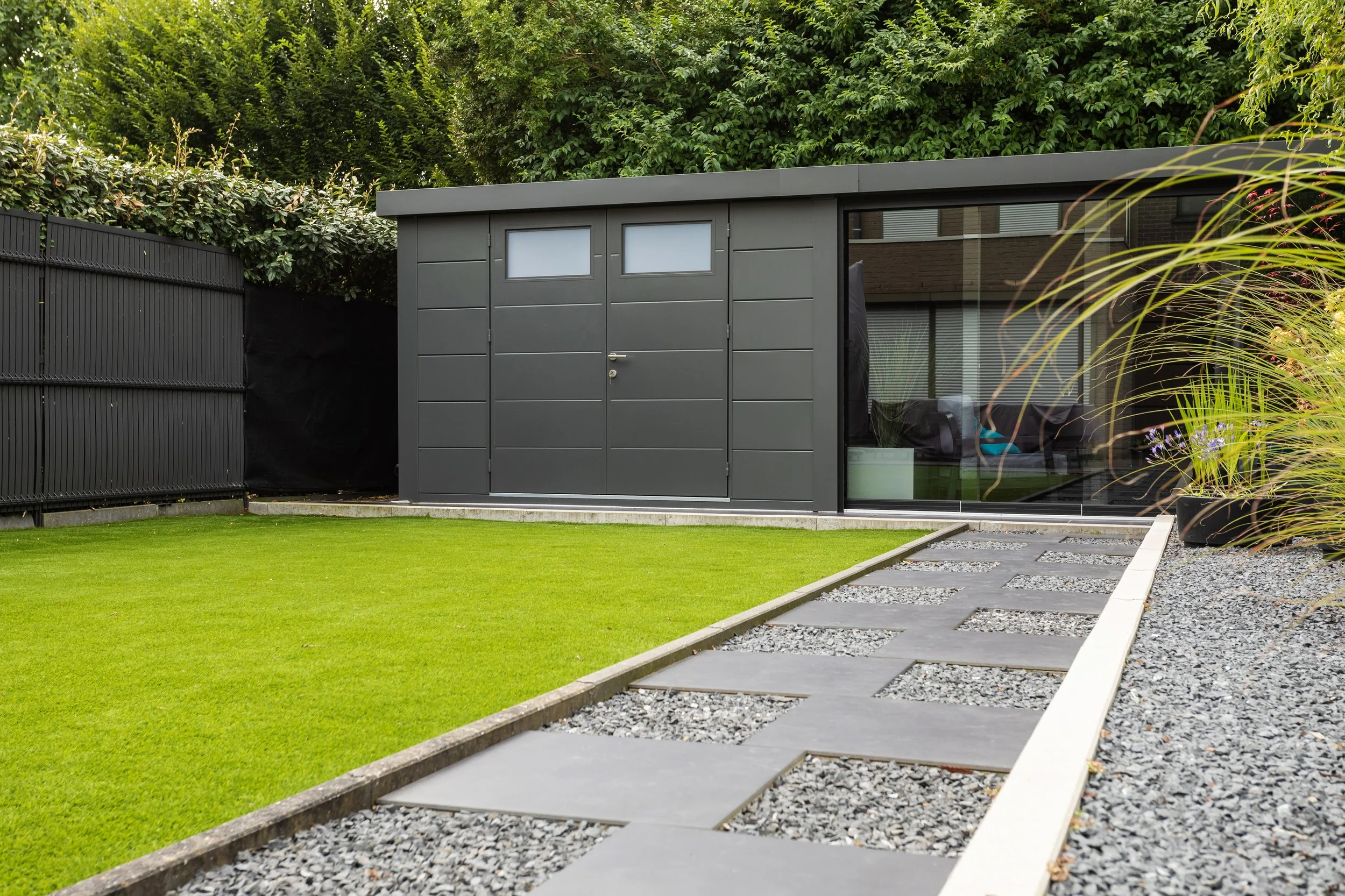 Modern gray shed in a landscaped backyard with a stepping stone pathway, a well-maintained lawn, and lush green foliage in the background.
