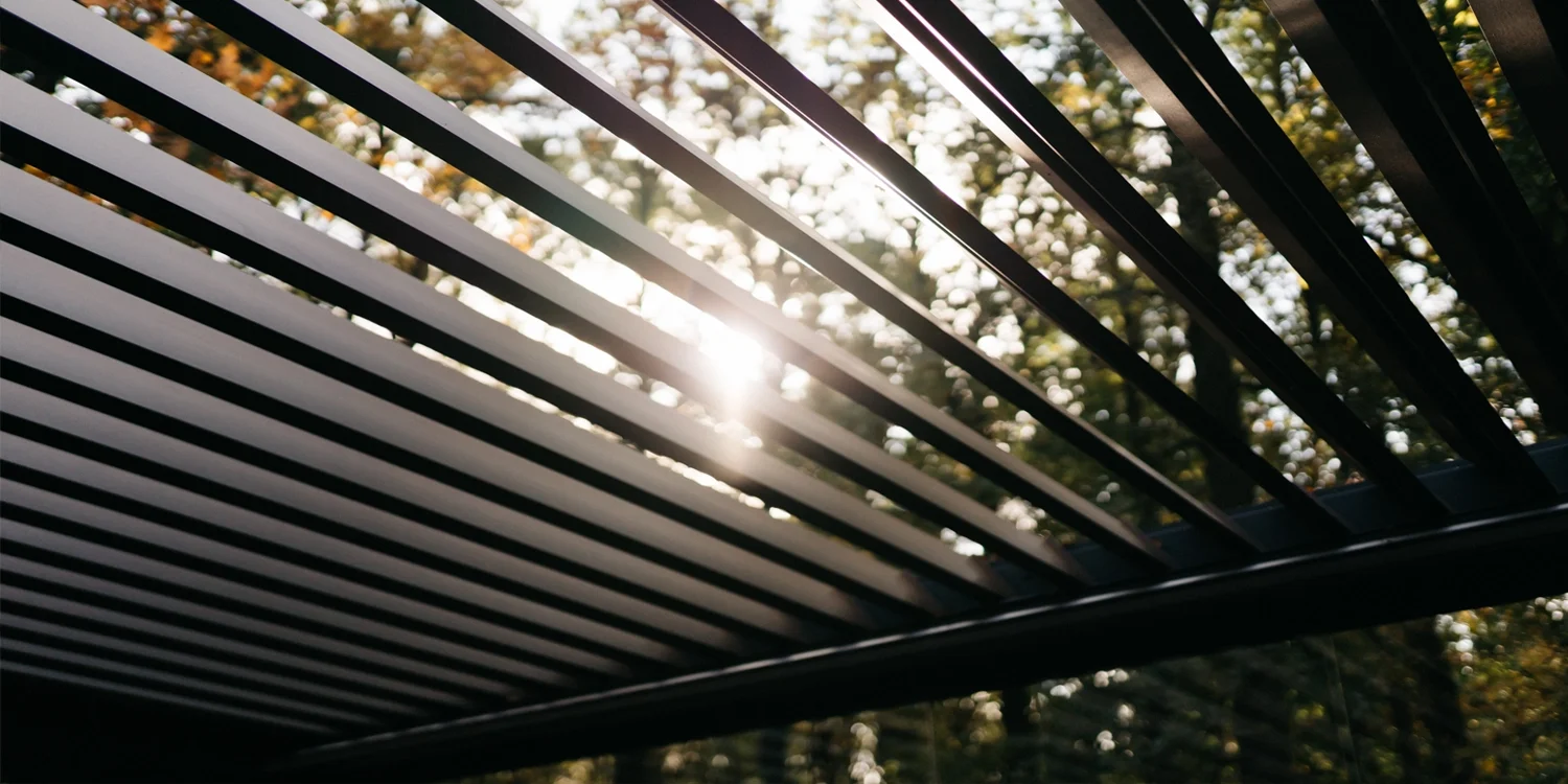 Close-up of a black metal outdoor grill with slats, sunlight shining through, blurred trees in the background.