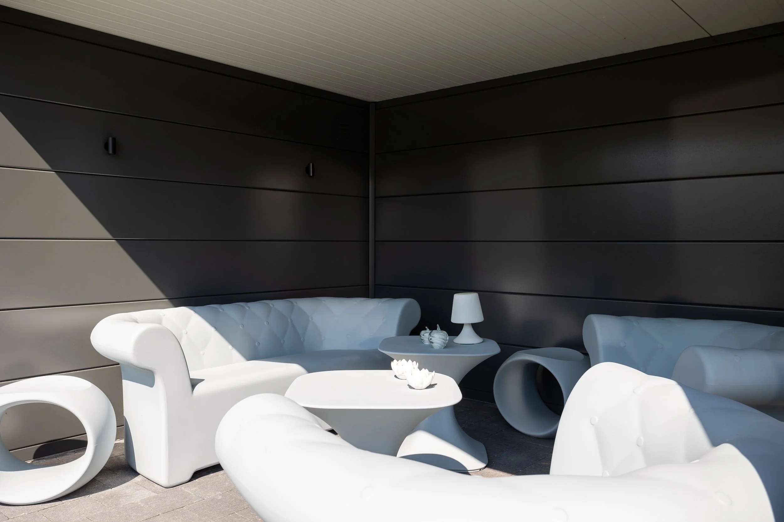 Outdoor seating area with white modern furniture, including armchairs, a side table, a small coffee table, and a table lamp, against a dark gray wall under a light-colored ceiling.