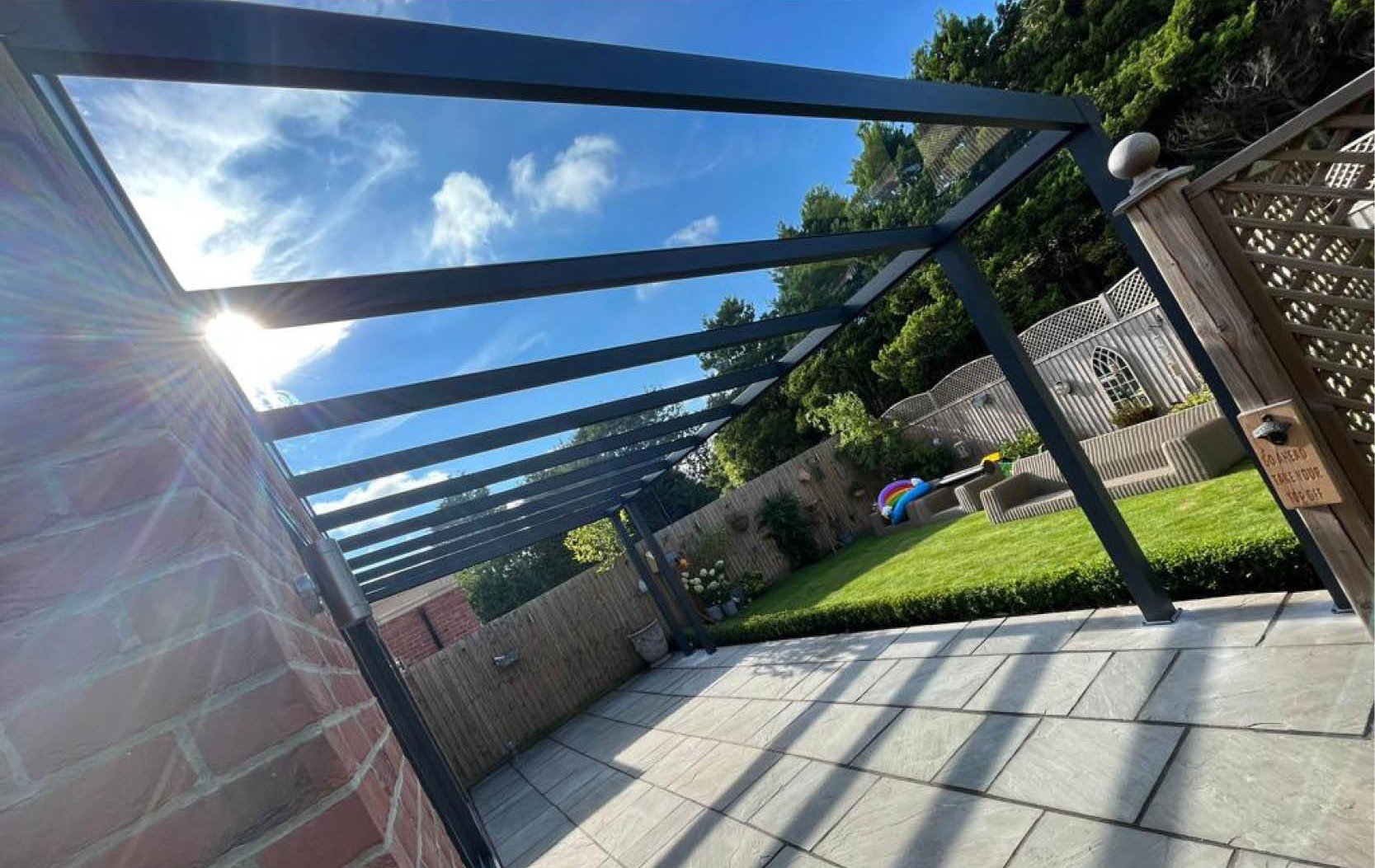 Backyard patio with a pergola, outdoor furniture, a grassy area with a trampoline, and a wooden fence with trees in the background.