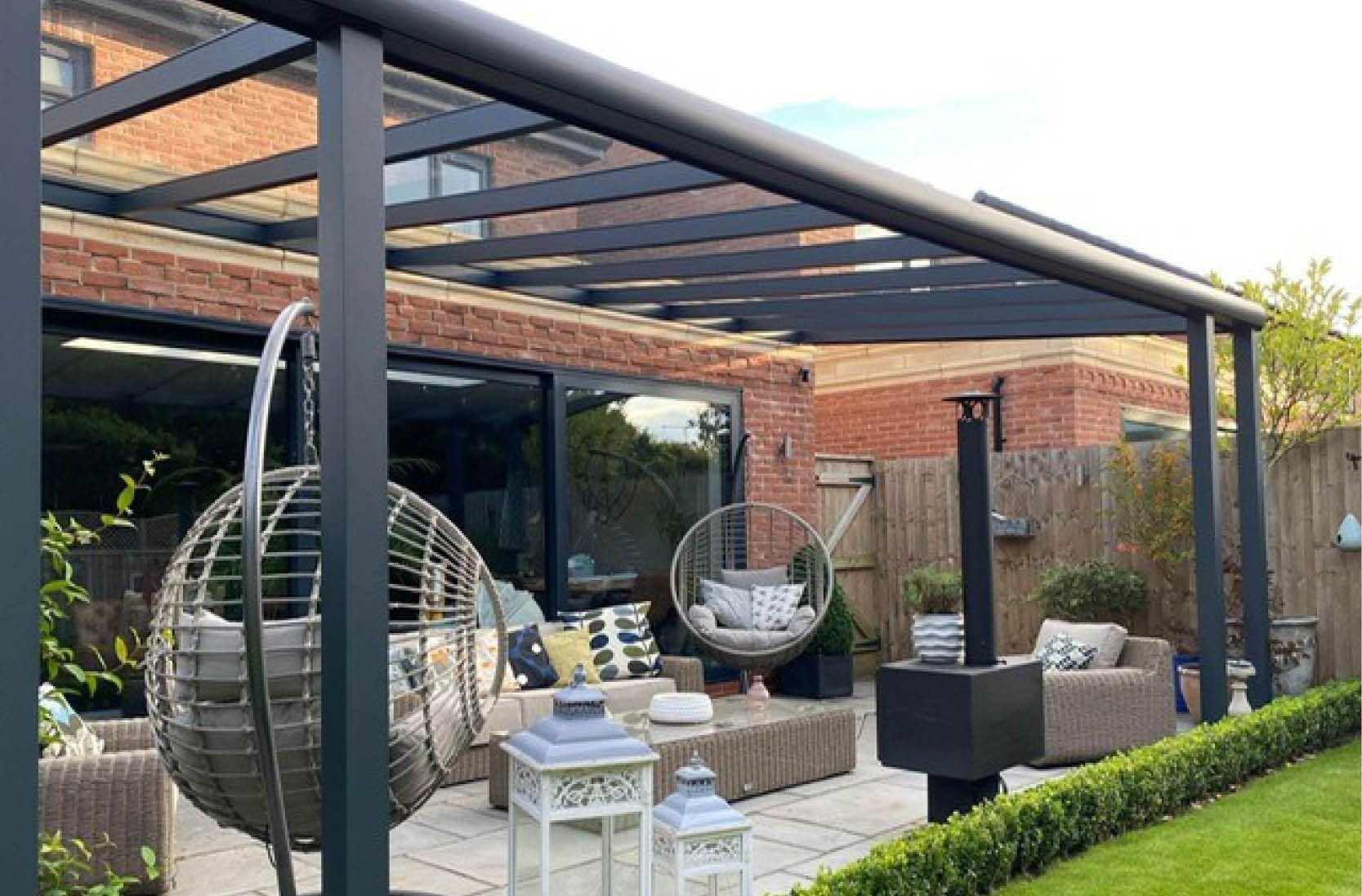Outdoor patio area with a dark metal pergola, wicker furniture with cushions, hanging egg chairs, potted plants, and a wooden fence, neighboring brick house, lush green lawn in foreground.