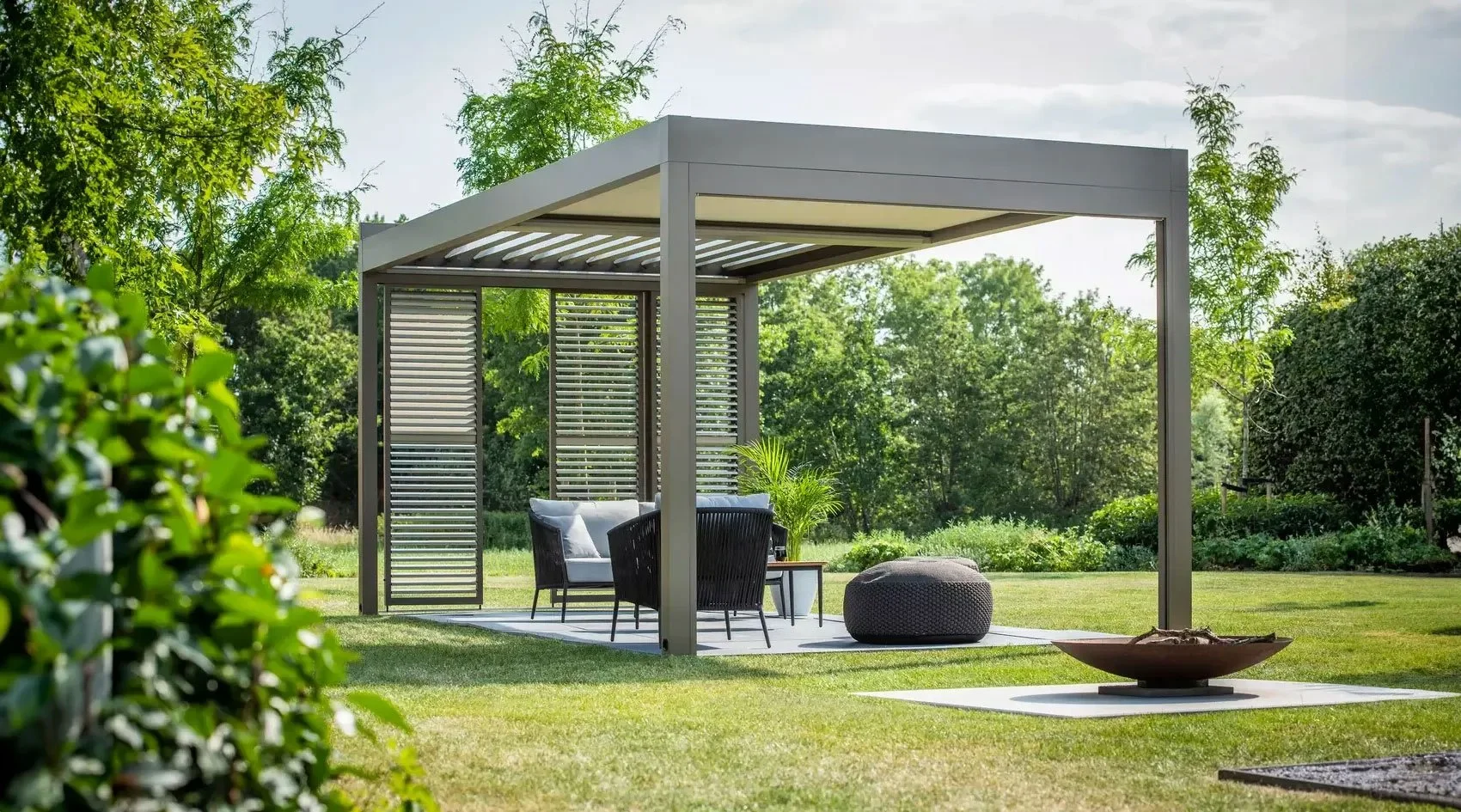 Outdoor patio area with a modern pergola, black chairs, a small table, a gray ottoman, and a fire pit, surrounded by lush green trees and grass.