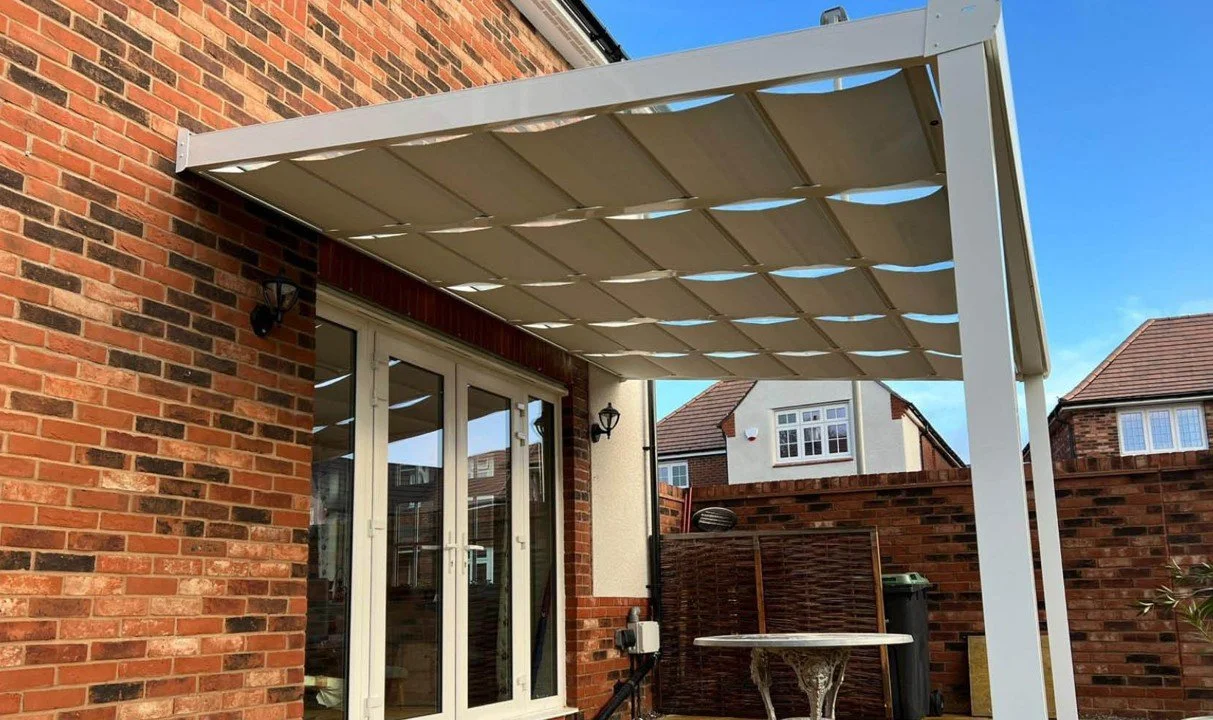 Photo of a backyard patio area featuring a white cover attached to a brick house wall, with double glass doors leading inside. There is a small round table with a decorative base and a black trash bin on the right. A brick privacy fence surrounds the