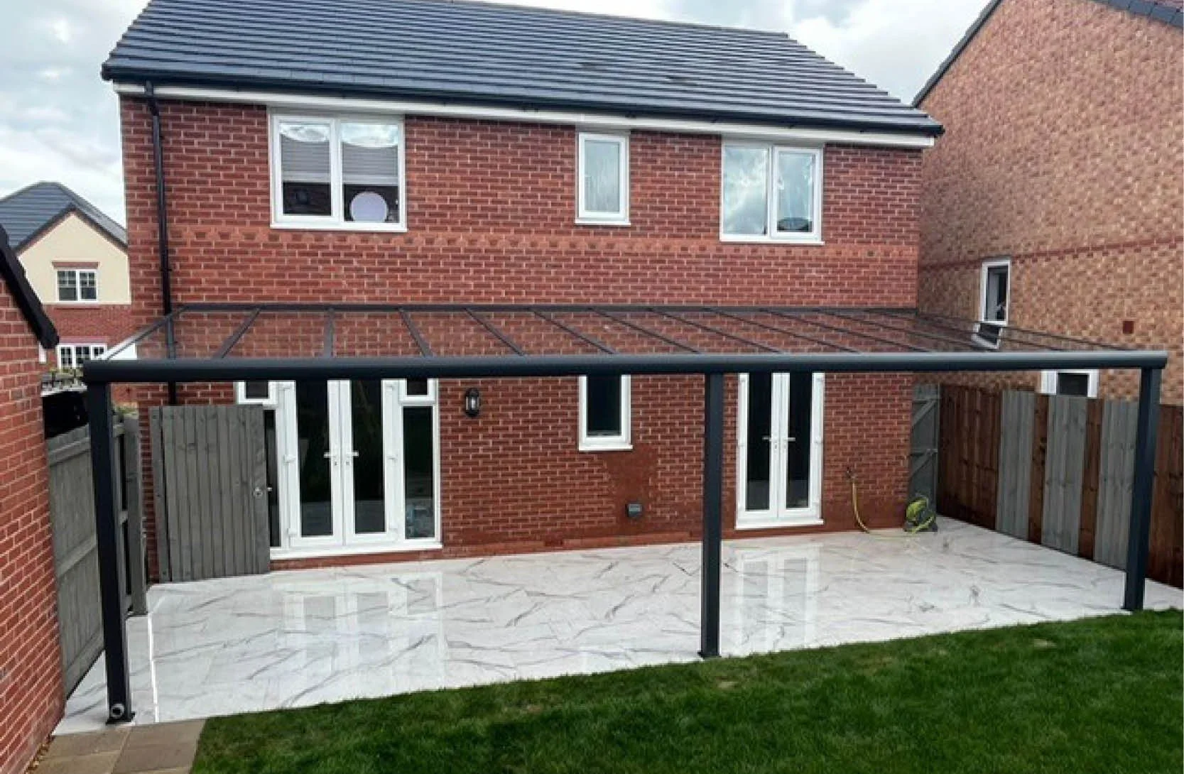 Backyard patio with white marble tiles, enclosed by brick and wooden fences, featuring a black metal and glass frame structure for a patio cover, attached to a two-story red brick house with multiple windows and a glass door.