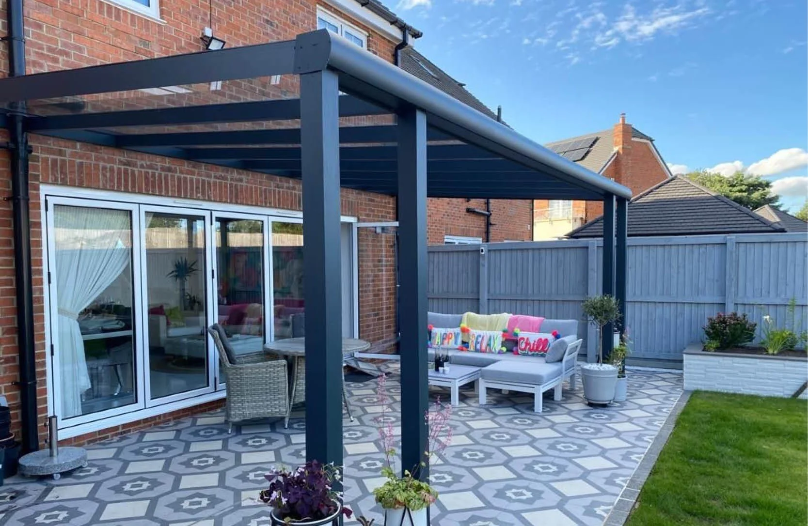 A backyard patio with patterned tile flooring, a white outdoor sofa with colorful pillows, potted plants, and a blue pergola structure. There is a brick house with large glass sliding doors, a nearby wooden fence, and a partly cloudy sky.
