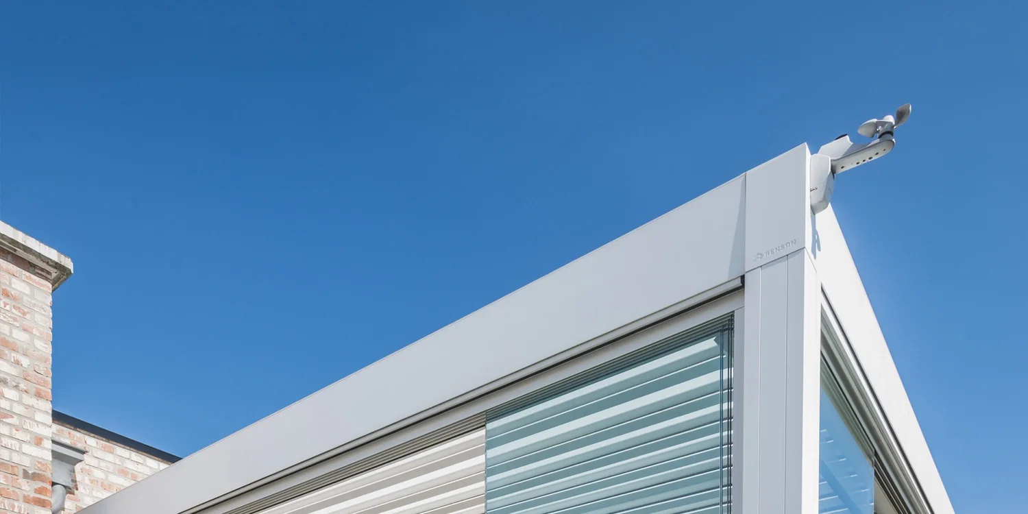 Close-up of a modern building corner with a white frame, glass windows with blinds, and a security camera at the top right against a clear blue sky.