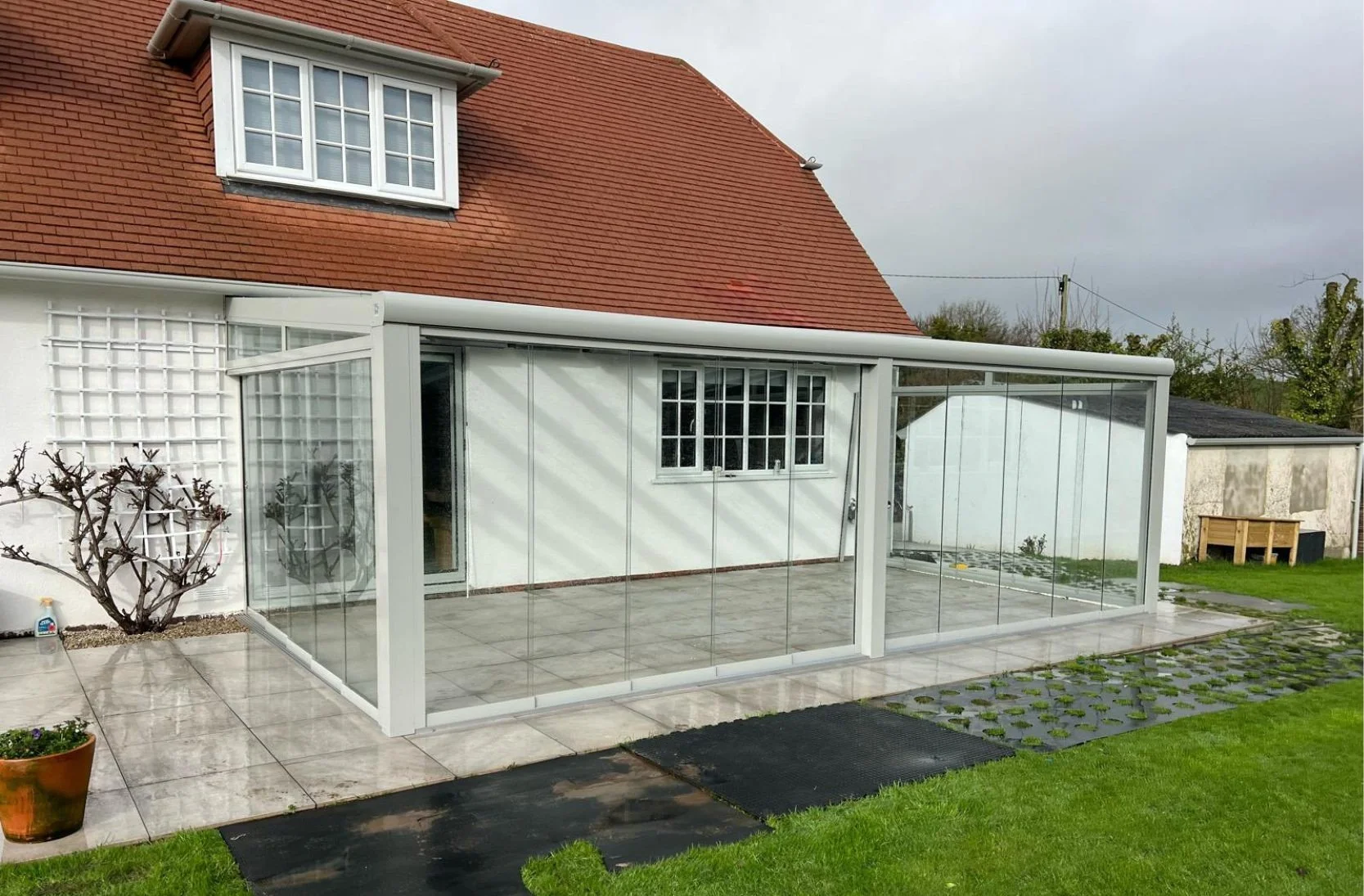 A backyard patio with a glass-enclosed sunroom attached to a house with a red tile roof. The patio has large tiled flooring, a potted plant, and a bare shrub. The surrounding lawn is green, and a small shed or garage is visible in the background. The