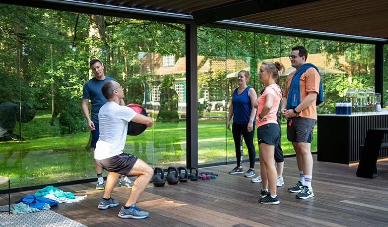 An outdoor fitness class with six people, one man holding a medicine ball while kneeling, demonstrating an exercise. Four participants stand in front of him, watching attentively, on a wooden deck with kettlebells and water bottles nearby. Large glass windows reveal a green backyard with trees and buildings.