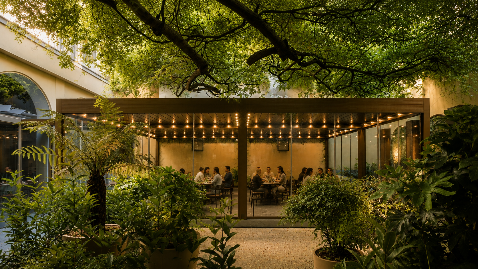 An outdoor dining area enclosed by glass walls, surrounded by lush green plants and trees, with people seated at tables inside. String lights decorate the ceiling, creating a warm ambiance.