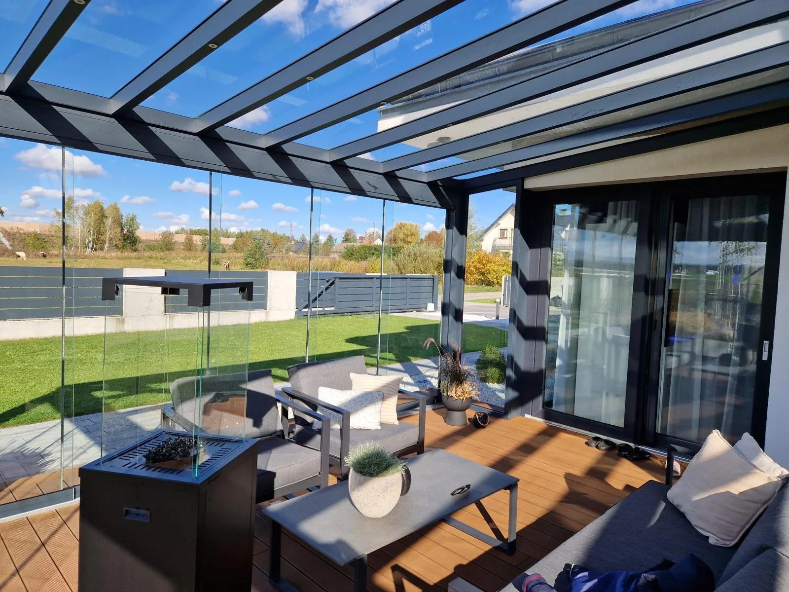 A modern enclosed patio with glass walls and a glass ceiling, furnished with outdoor seating, a small table with plants, and a view of a green yard with trees and a fence outside.