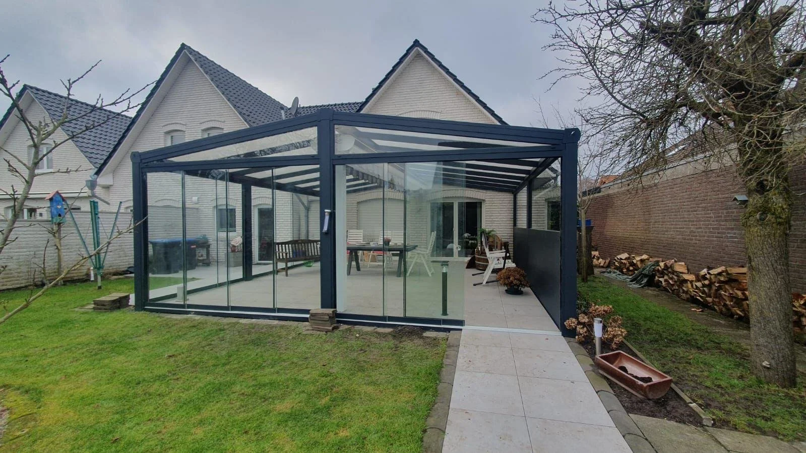 A modern glass-enclosed patio with outdoor furniture in a backyard, surrounded by a grassy lawn and neighboring houses.