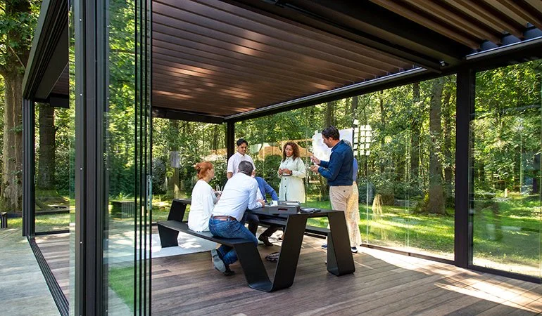 A group of people in a modern glass conference room surrounded by trees, engaged in a meeting or discussion.