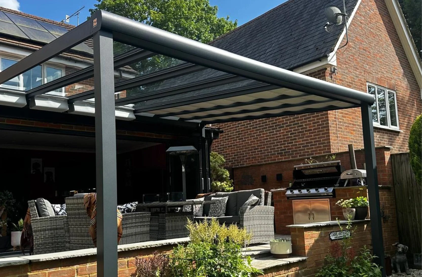 Outdoor patio with wicker furniture, a barbecue grill, and a covered outdoor area attached to a brick house with a window and lantern. Green plants and trees are visible in the background.