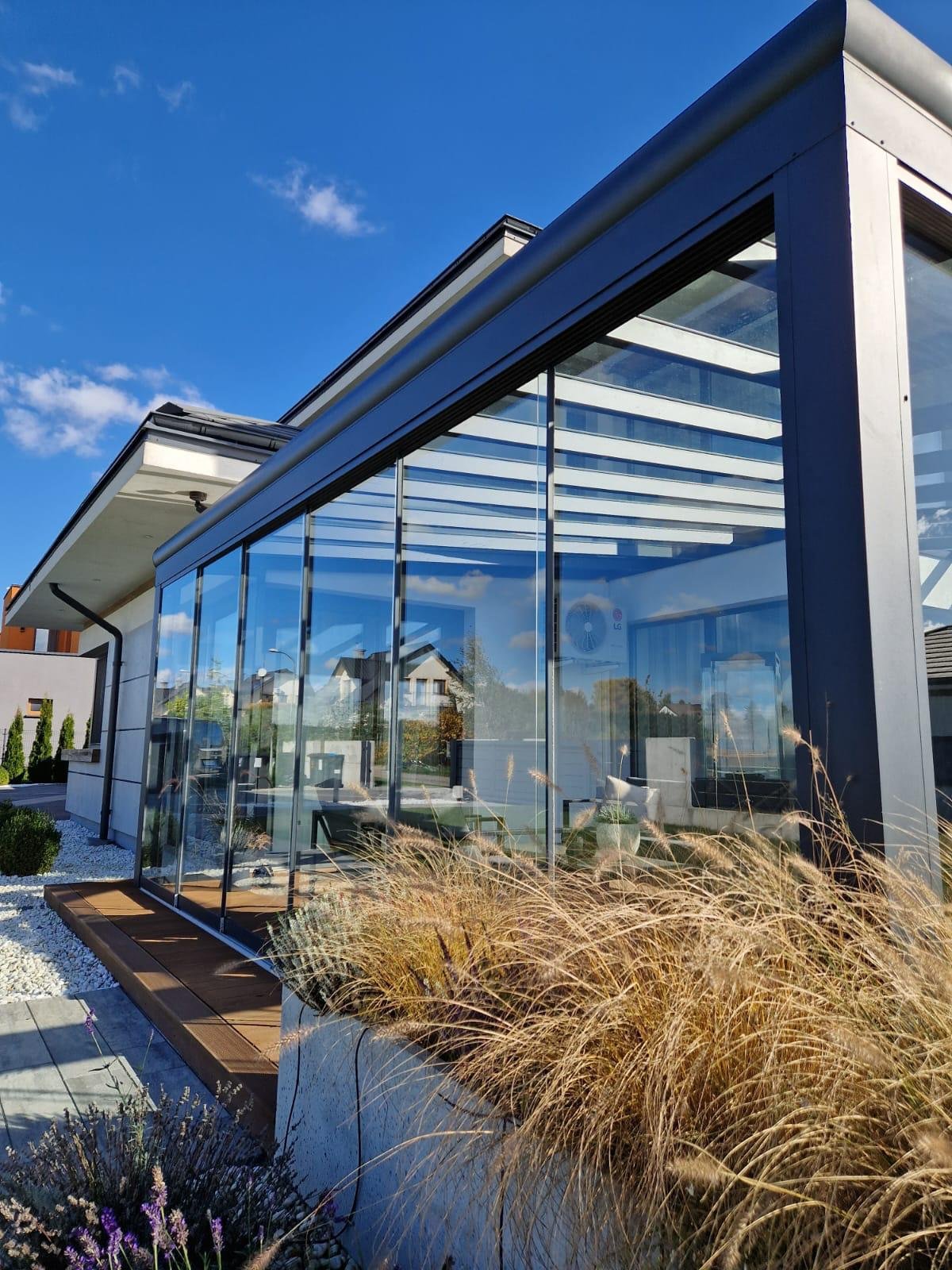 Modern house with large glass windows and a small wooden deck, surrounded by ornamental grass and flowering plants, under a blue sky with a few clouds.