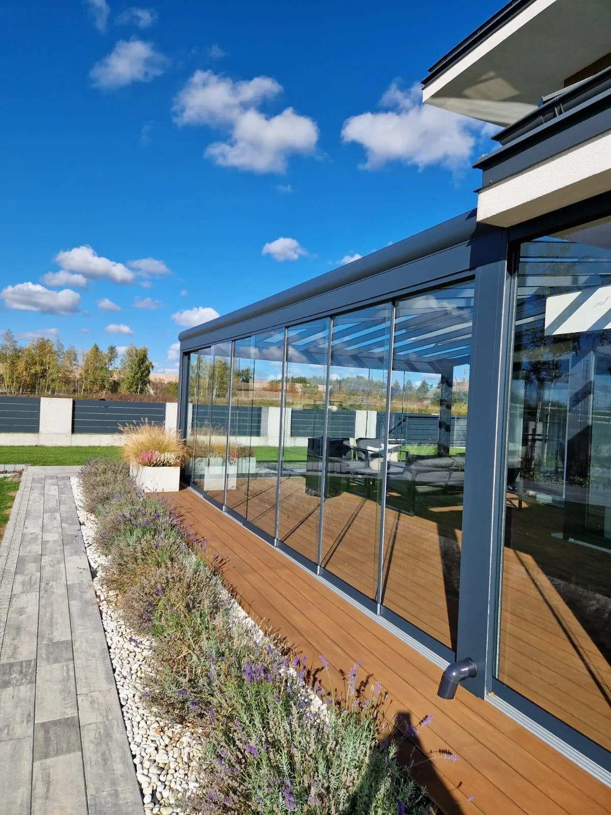 A modern house with a glass sunroom, outdoor wooden deck, garden with lavender plants, and a bright blue sky with scattered clouds.