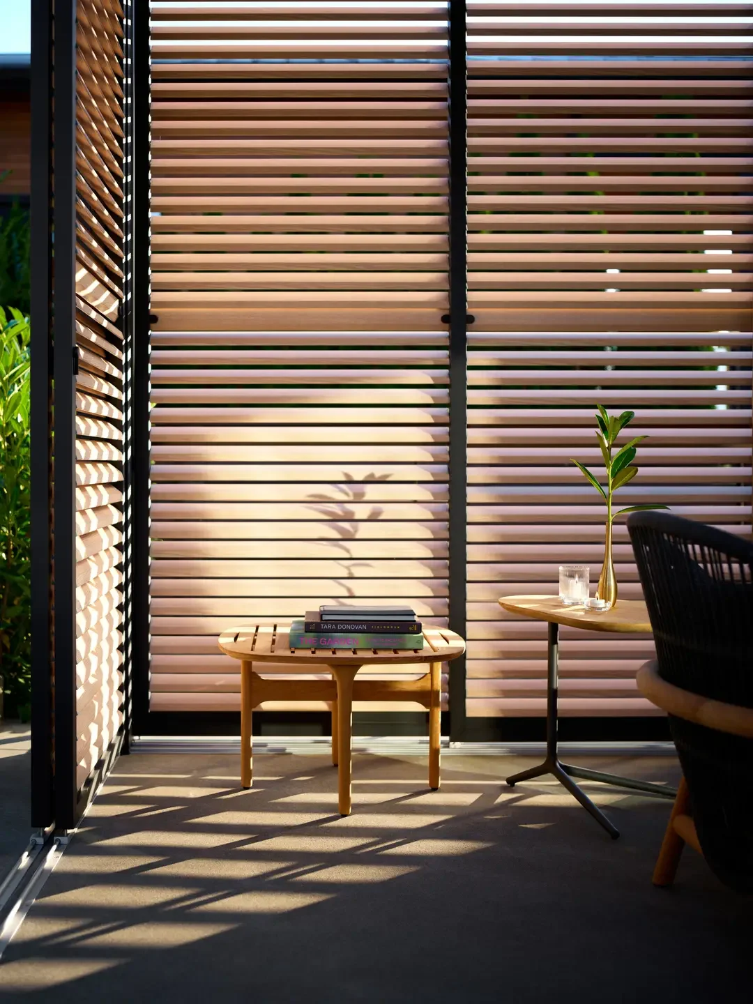 A cozy outdoor sitting area with a small wooden table, books on top, a round side table with a plant in a gold vase, and a glass of water. The area is partially shaded by a striped privacy screen, casting shadows on the concrete floor.