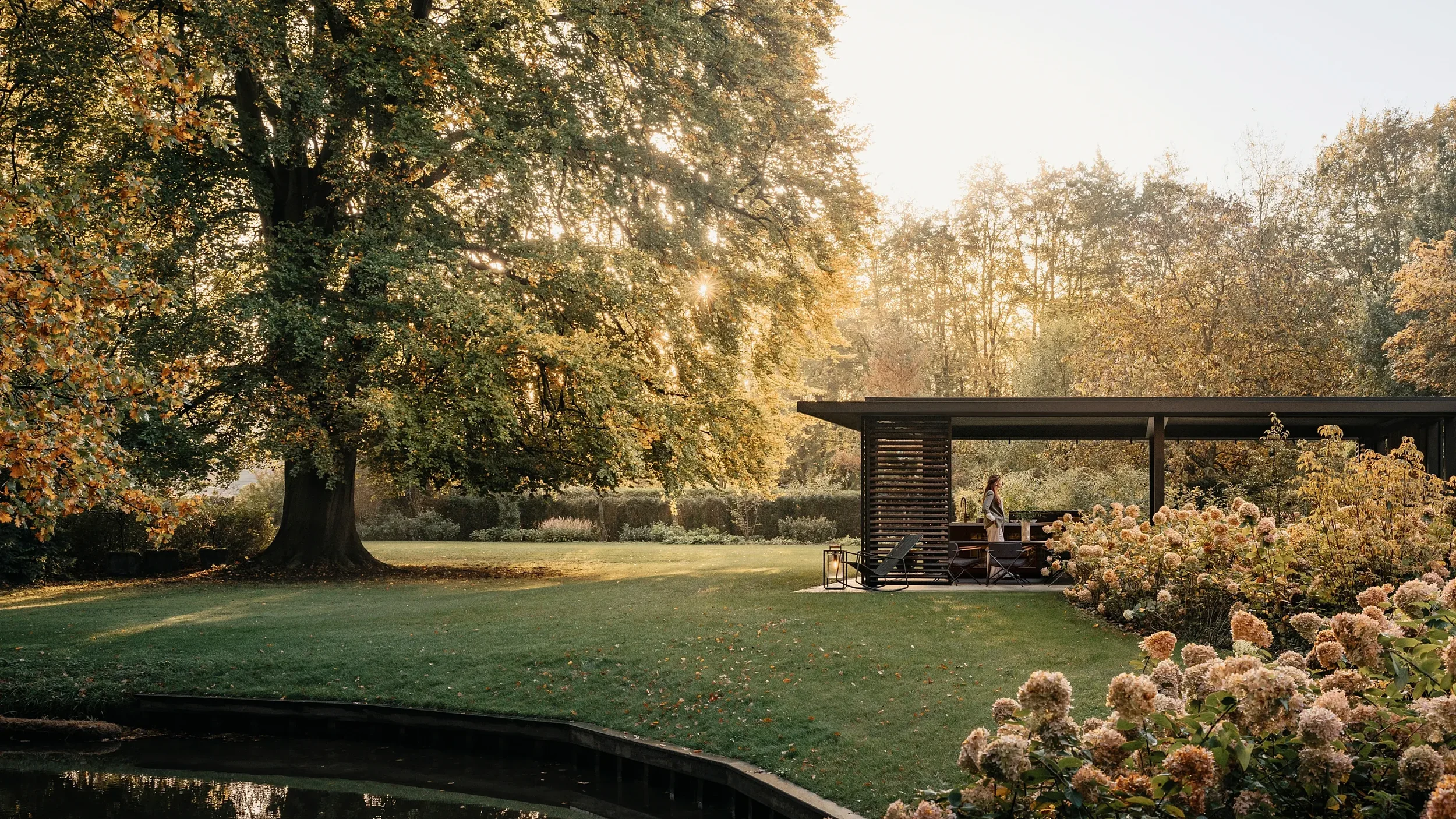 A woman stands near a modern outdoor pavilion in a garden at sunset, surrounded by large trees with autumn foliage and blooming flowers.
