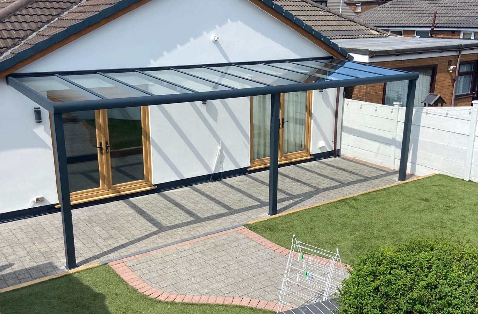 Backyard with a paved patio, a white raised lawn, a white fence, and a glass-top patio cover attached to a house with white walls and wooden-framed sliding doors.