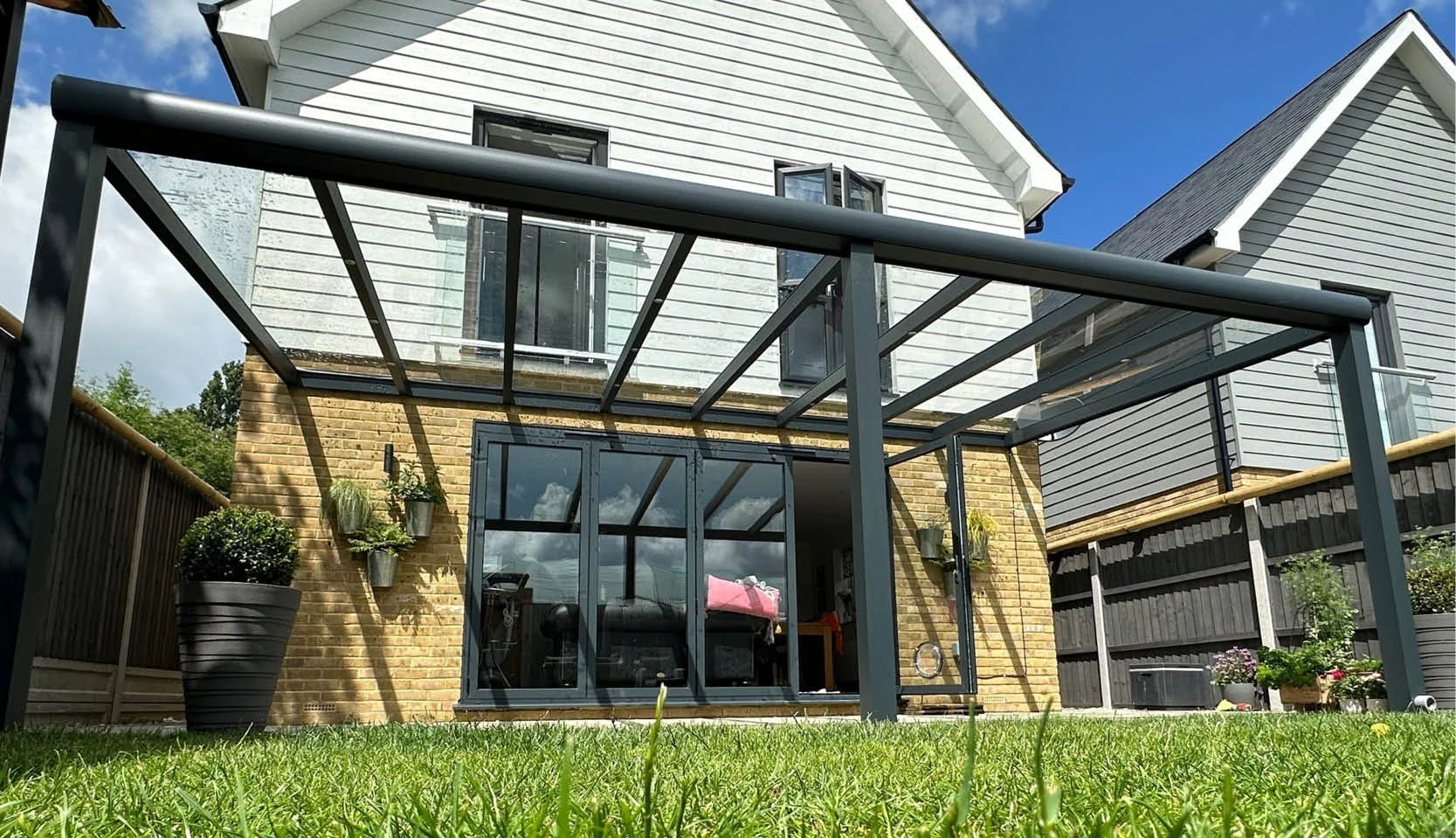 Backyard with a glass-roofed patio, potted plants, a lawn, and a two-story house with white siding and black window frames.
