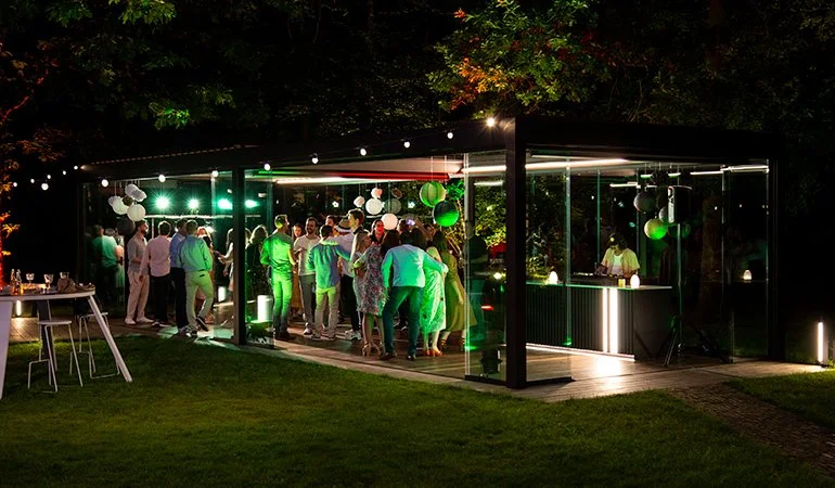 At night, people gather inside a glass-walled party pavilion decorated with hanging green and white lights and spheres. There are trees in the background, and a table with chairs is outside on the grass.
