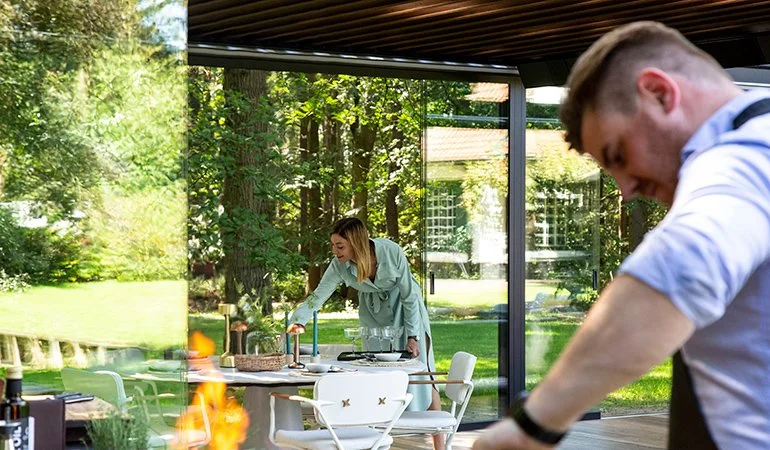 A woman and a man are cooking outside on a patio with a wooded backyard in the background. The woman is leaning over a table near a grill, while the man is focused on a task in the foreground.