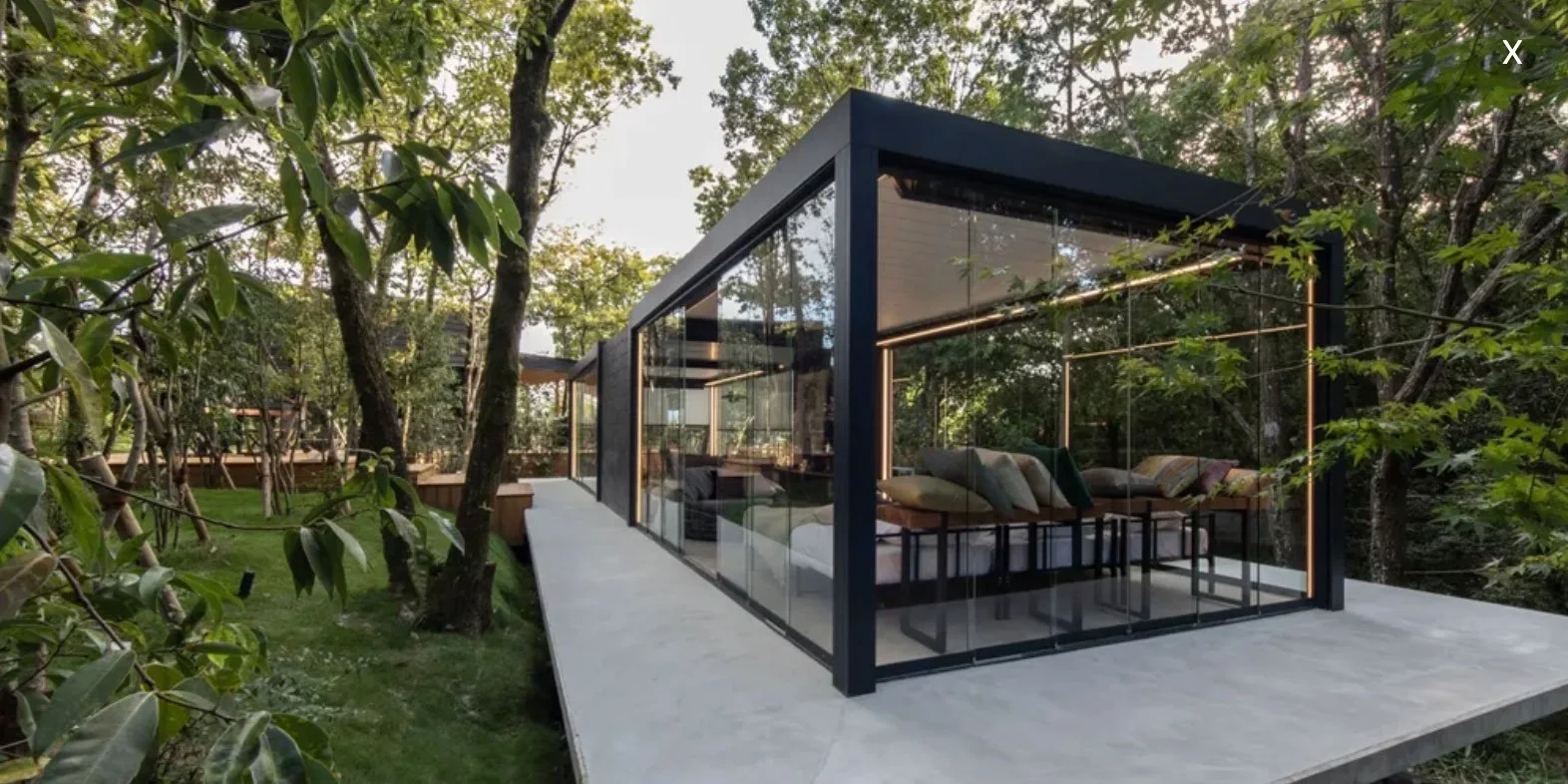 Modern glass house surrounded by trees, with a concrete deck extending from it.