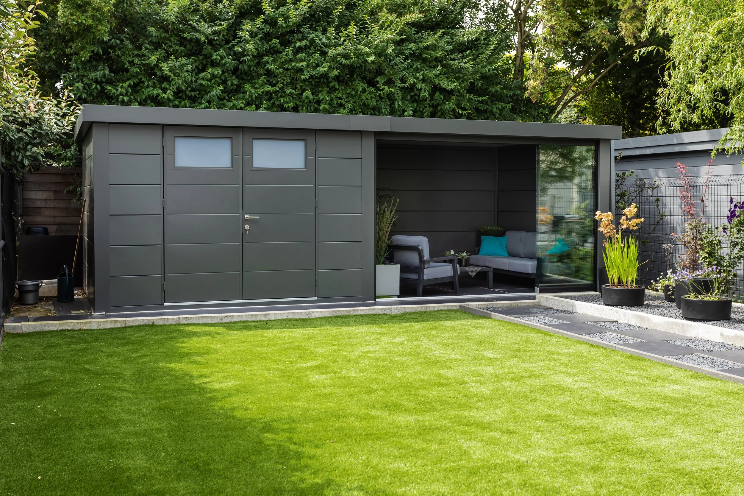 Modern backyard with a green lawn, gray shed, and a covered seating area with gray sofa and chairs, surrounded by potted plants and trees.