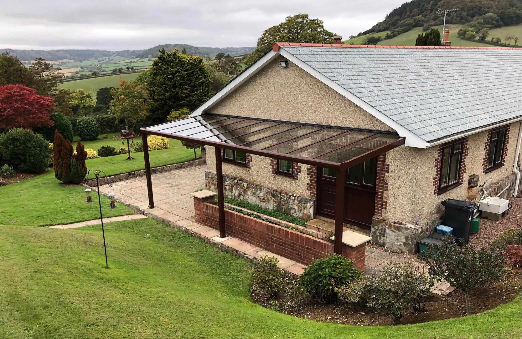 A house with a stone and wood exterior, featuring a small porch with a transparent roof, is surrounded by a well-maintained garden with green grass, shrubs, and trees. In the background, rolling hills and a cloudy sky are visible.