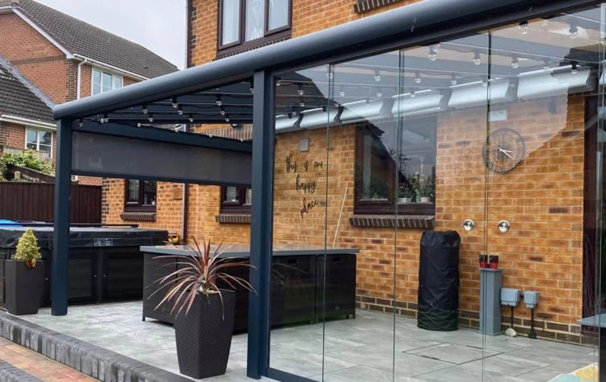 Modern outdoor patio with glass enclosure, potted plants, brick house backdrop, and string lights.