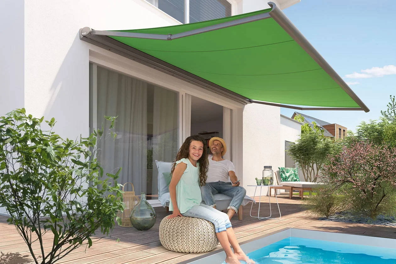 A woman and a girl sitting on a pouf and a lounge chair on a wooden patio next to a small swimming pool, with a green retractable awning above them, surrounded by plants and a modern house in the background.