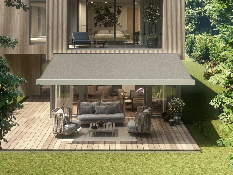 Modern house with a wooden deck and an outdoor patio area featuring grey wicker furniture, a sofa, two armchairs with cushions, a coffee table, potted plants, and an awning, with a view of a bedroom upstairs with glass walls.