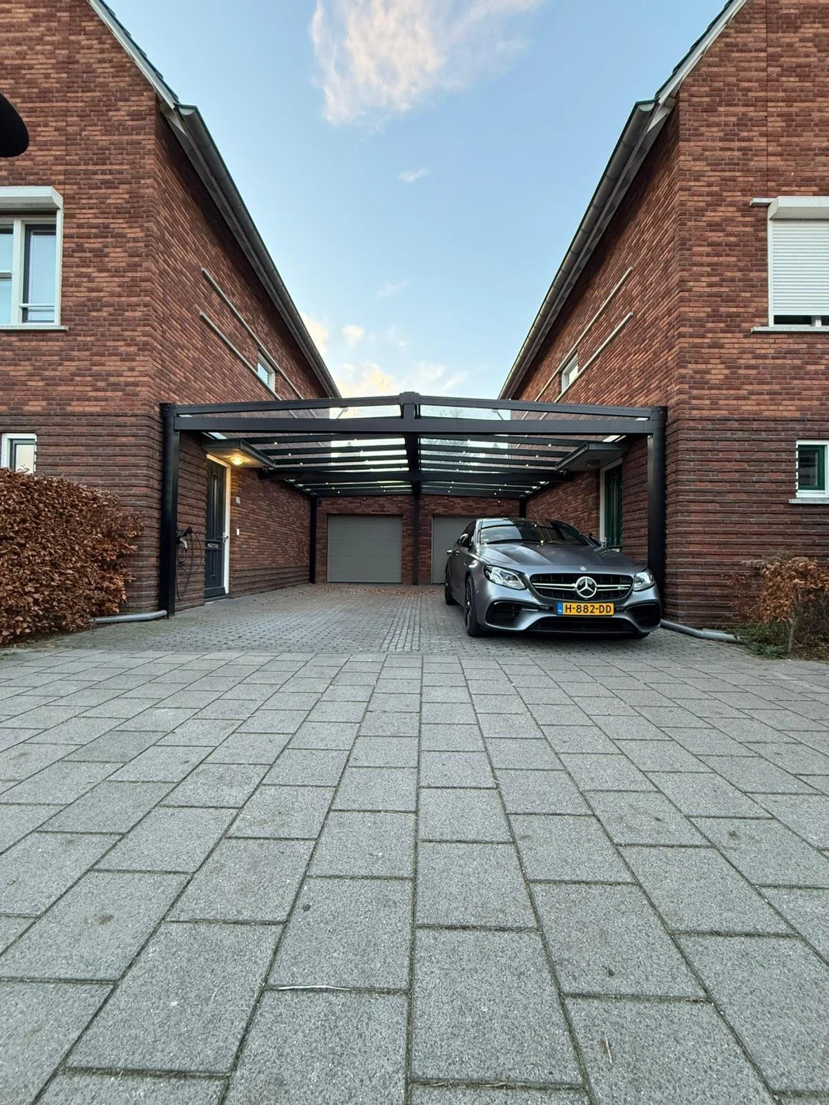 A modern residential driveway with a parked silver Mercedes-Benz car under a transparent carport, brick buildings on either side, and a paved surface in front with bushes on the sides.