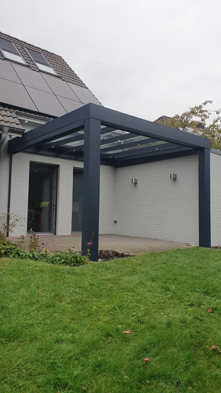 A modern house with solar panels on the roof and a clear glass patio cover supported by four dark blue pillars, with a small garden and grass in the foreground.