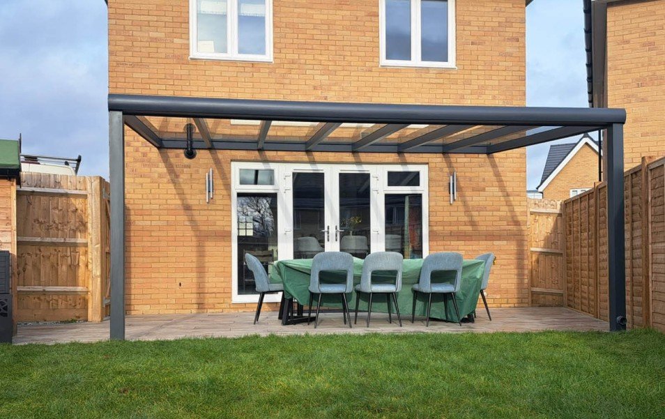 Backyard patio with a table covered by a green tablecloth and six gray chairs, a glass door leading inside, and a modern black metal pergola overhead.