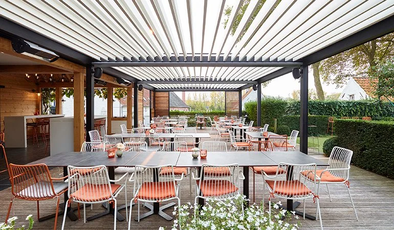 Outdoor dining area with tables and chairs under a pergola, surrounded by greenery.