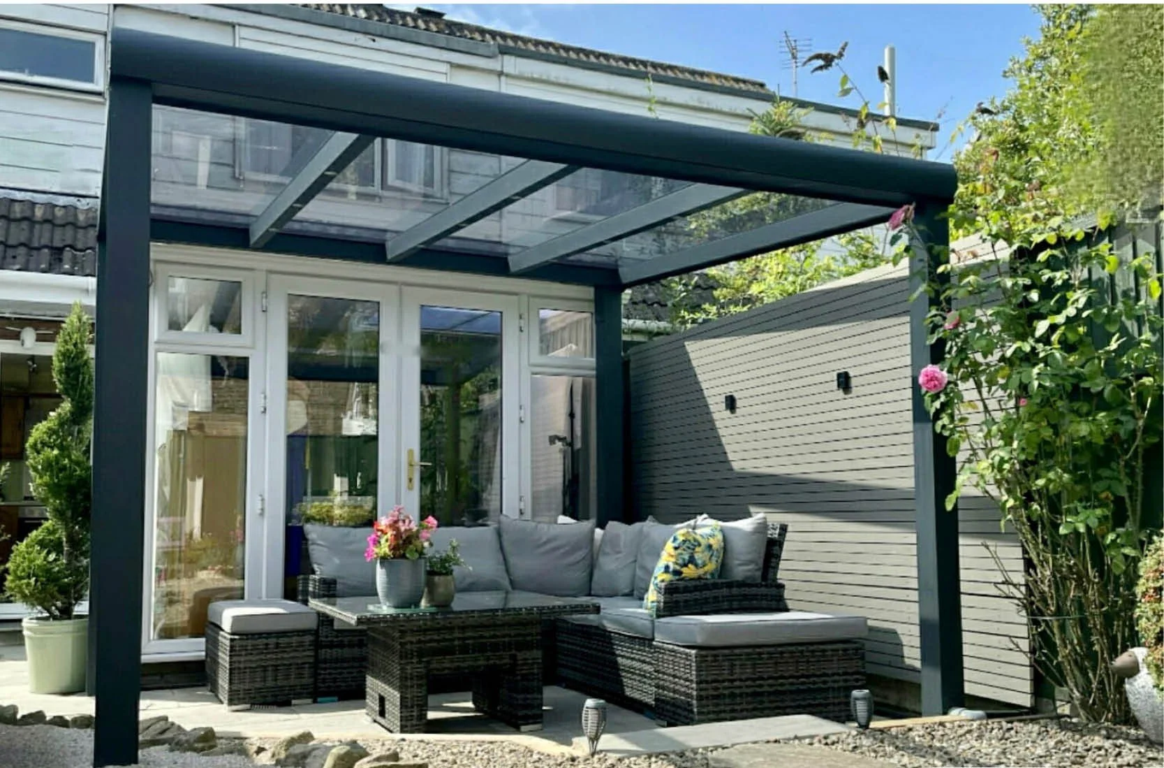 A backyard patio with a black wicker outdoor sofa with cushions, a glass-topped coffee table with potted flowers, and a pergola with a transparent roof. There is a door leading inside the house, a privacy fence, and greenery including flowering plant