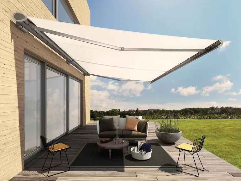 Outdoor patio with a modern sofa, two wire chairs with yellow cushions, a round coffee table, a potted plant, and a retractable white awning attached to a wooden house, overlooking a grassy field under a partly cloudy sky.