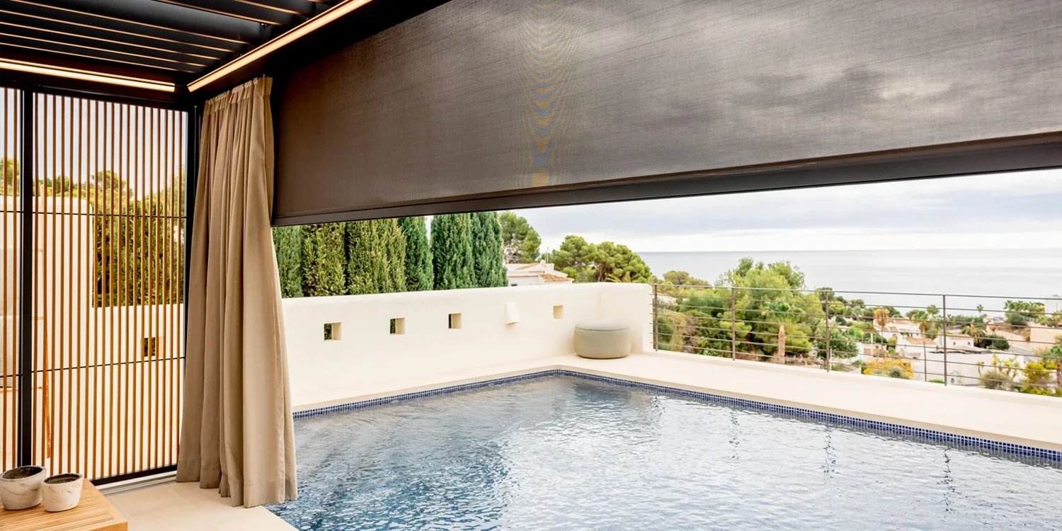 View of a private outdoor infinity pool with a scenic ocean view, surrounded by a white wall, greenery, and a partial view of a patio area.