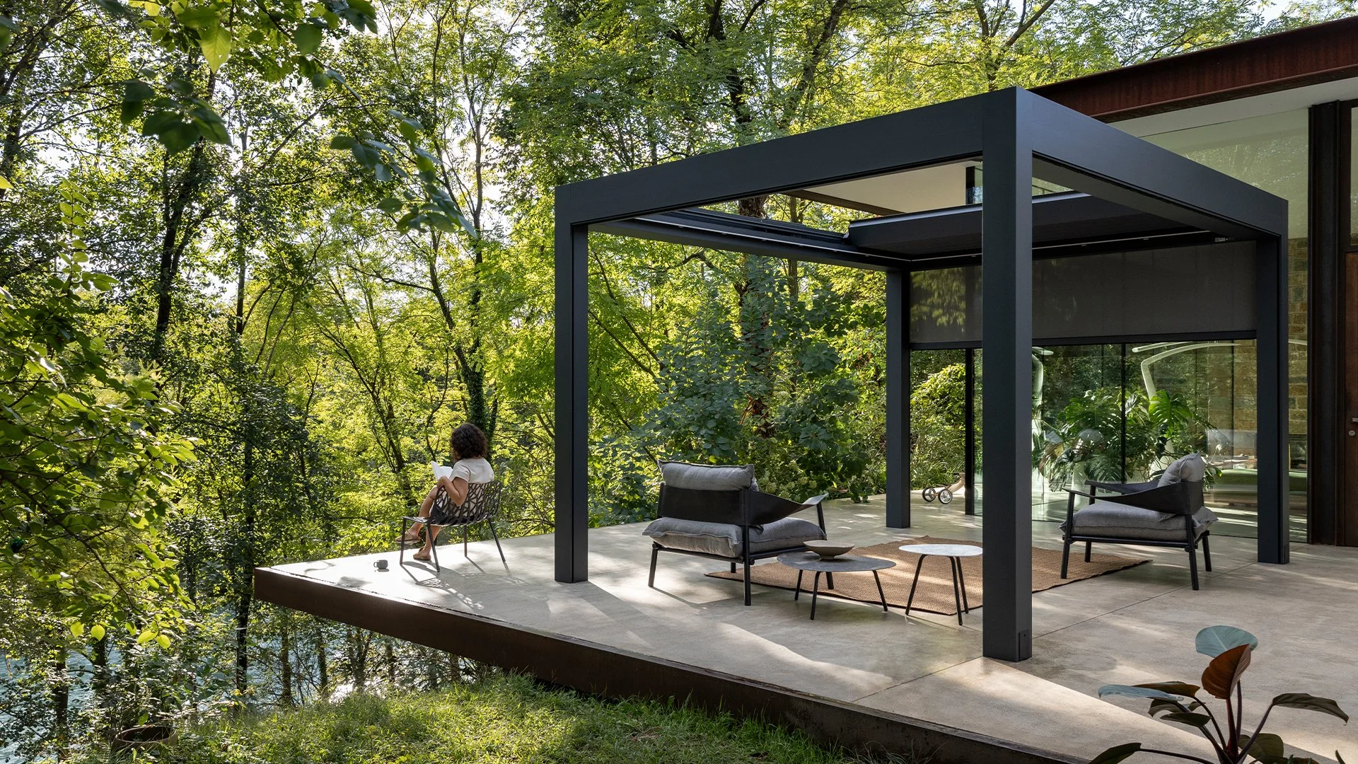 Modern outdoor patio with seating area overlooking a lush green forest, with a person sitting on a chair reading.