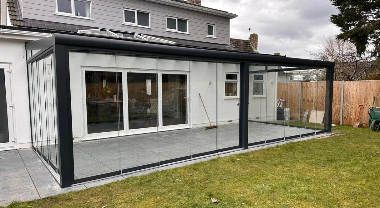 A backyard with a glass enclosed patio extension attached to a house, with sliding glass doors and tiled flooring, fenced with wood panels, and surrounded by a grassy lawn.