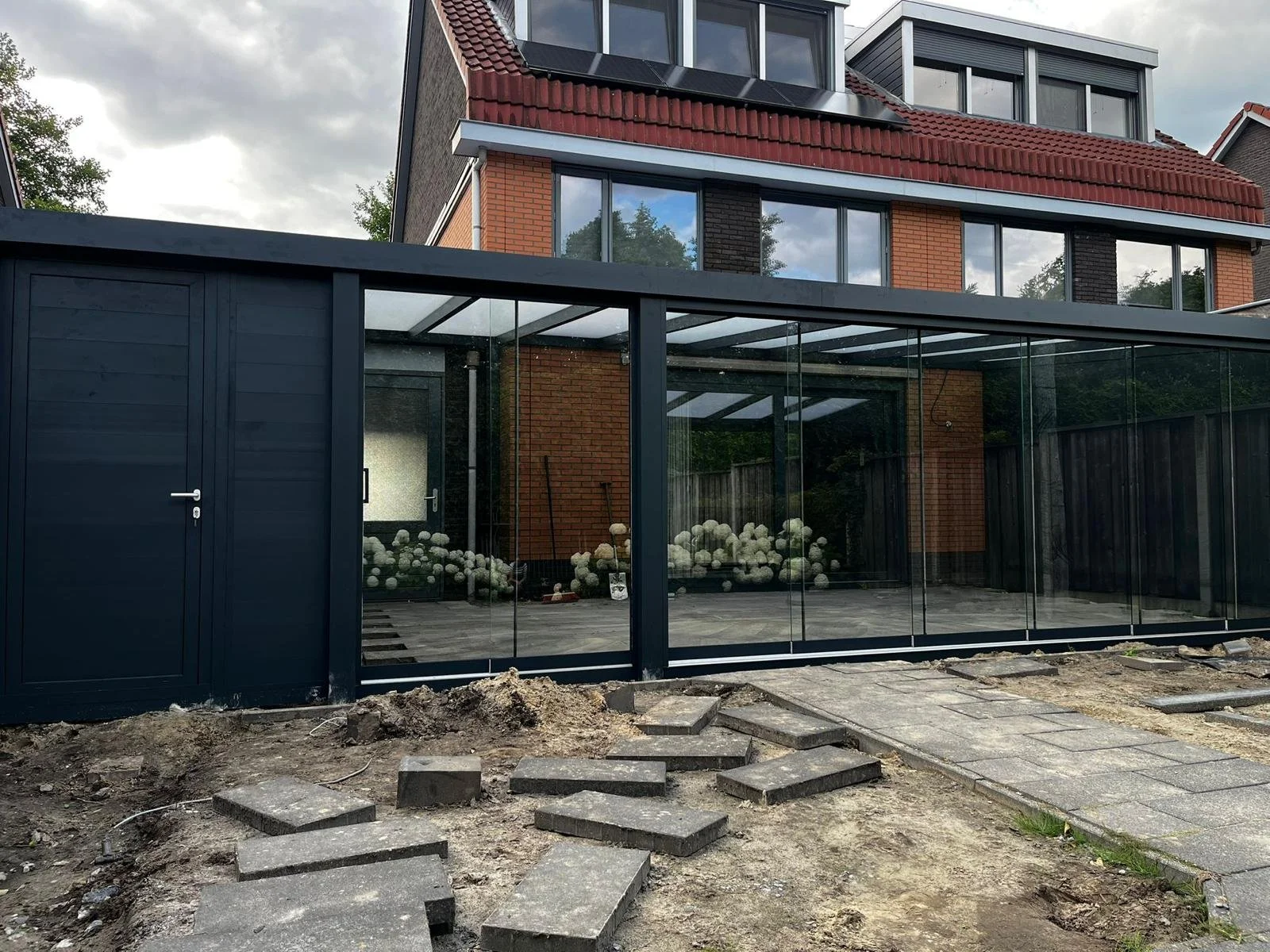 Update of a rear patio and conservatory on the back of a house with black-framed glass walls, brick and modern siding, surrounding a newly paved area with some construction debris and stepping stones.