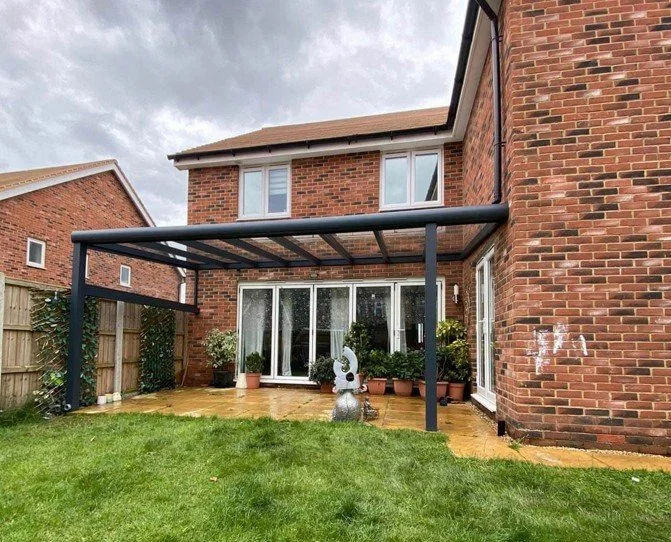 Backyard with a brick house, a patio, potted plants, and a black metal and glass patio cover on a cloudy day.