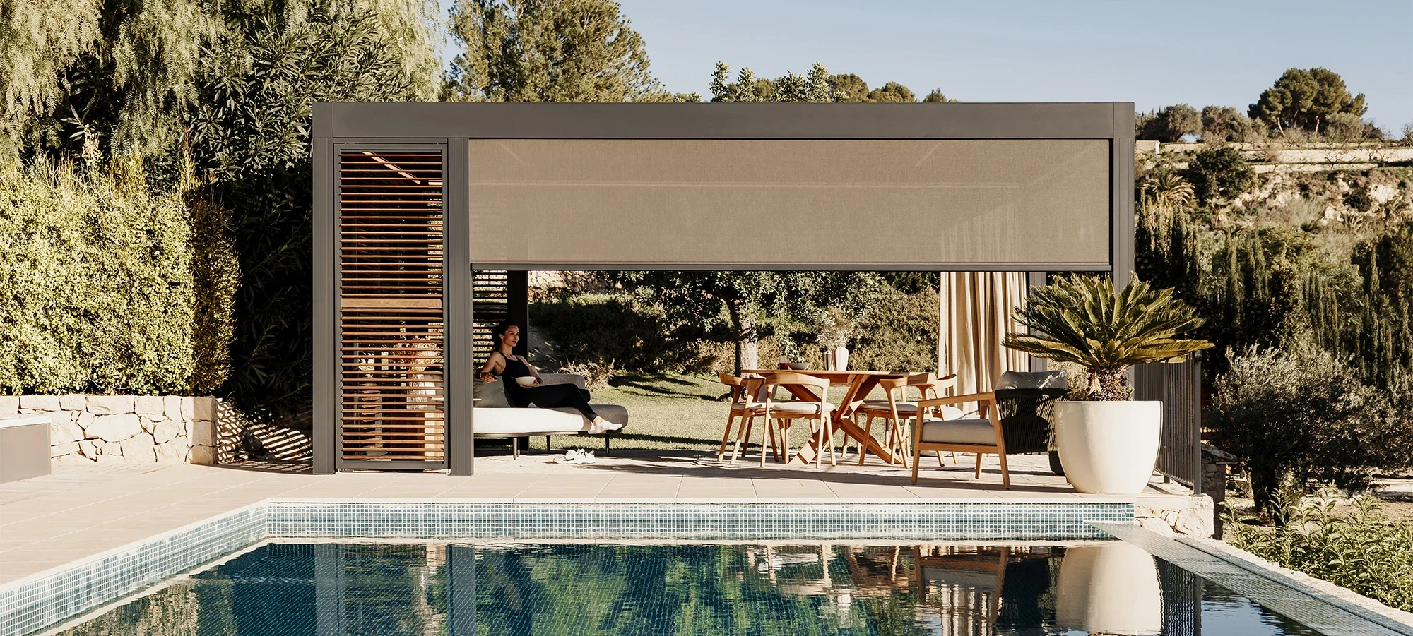 A modern outdoor patio area with a pool, shaded seating area with a woman sitting on a lounge chair holding a bowl, a wooden dining table with chairs, large white pot with a plant, and surrounded by greenery and trees.